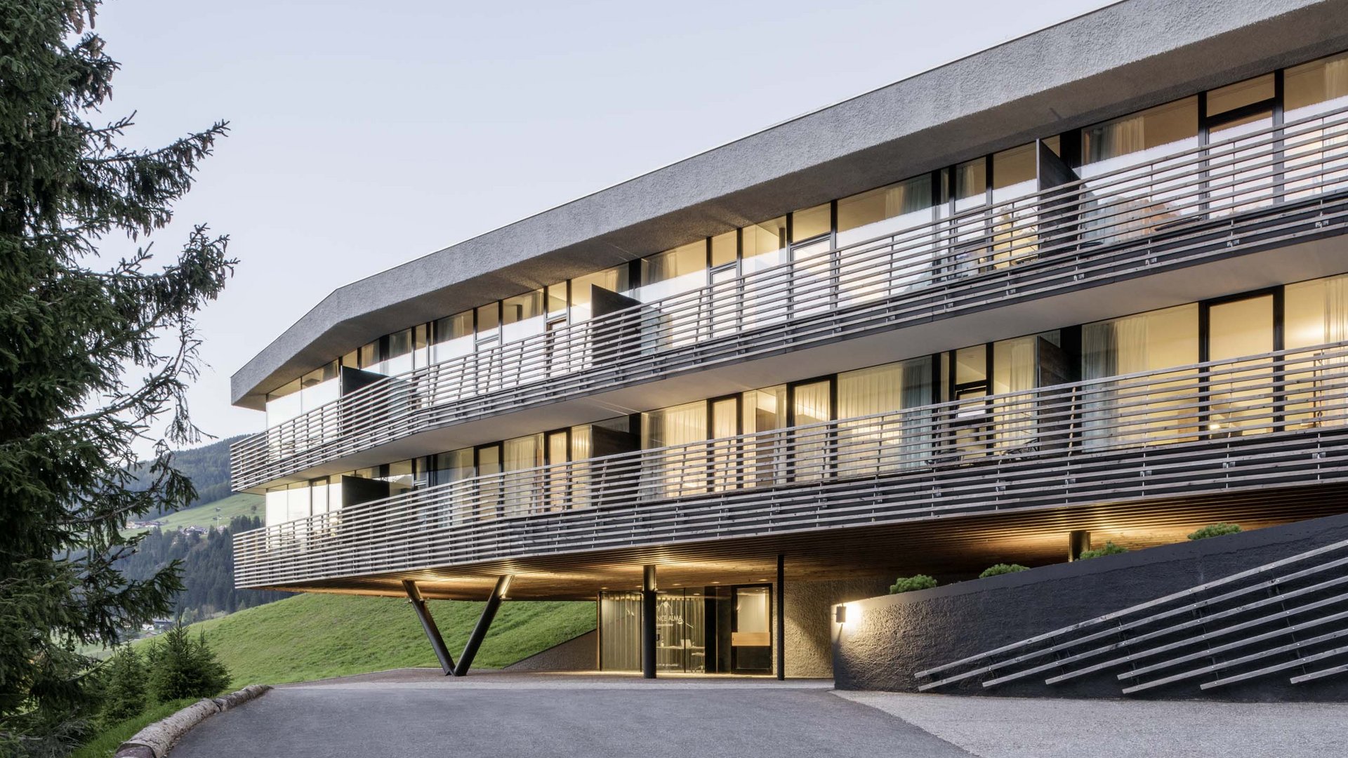 Urlaub mit Stil in der Residence Alma Paramout Modernes Hotelgebäude in den Bergen mit beleuchteten Fenstern bei Abenddämmerung