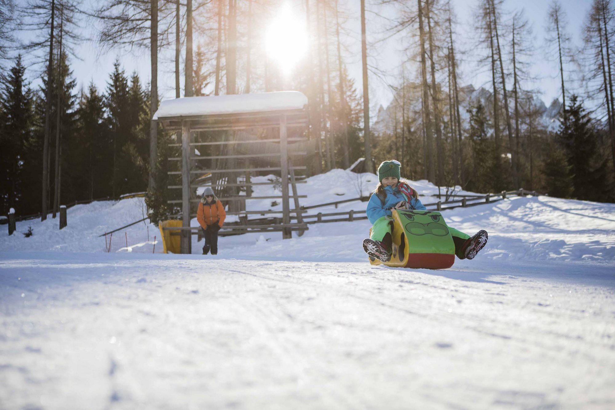 Our family experiences Child sledding on snow with colorful sled, adult standing in background
