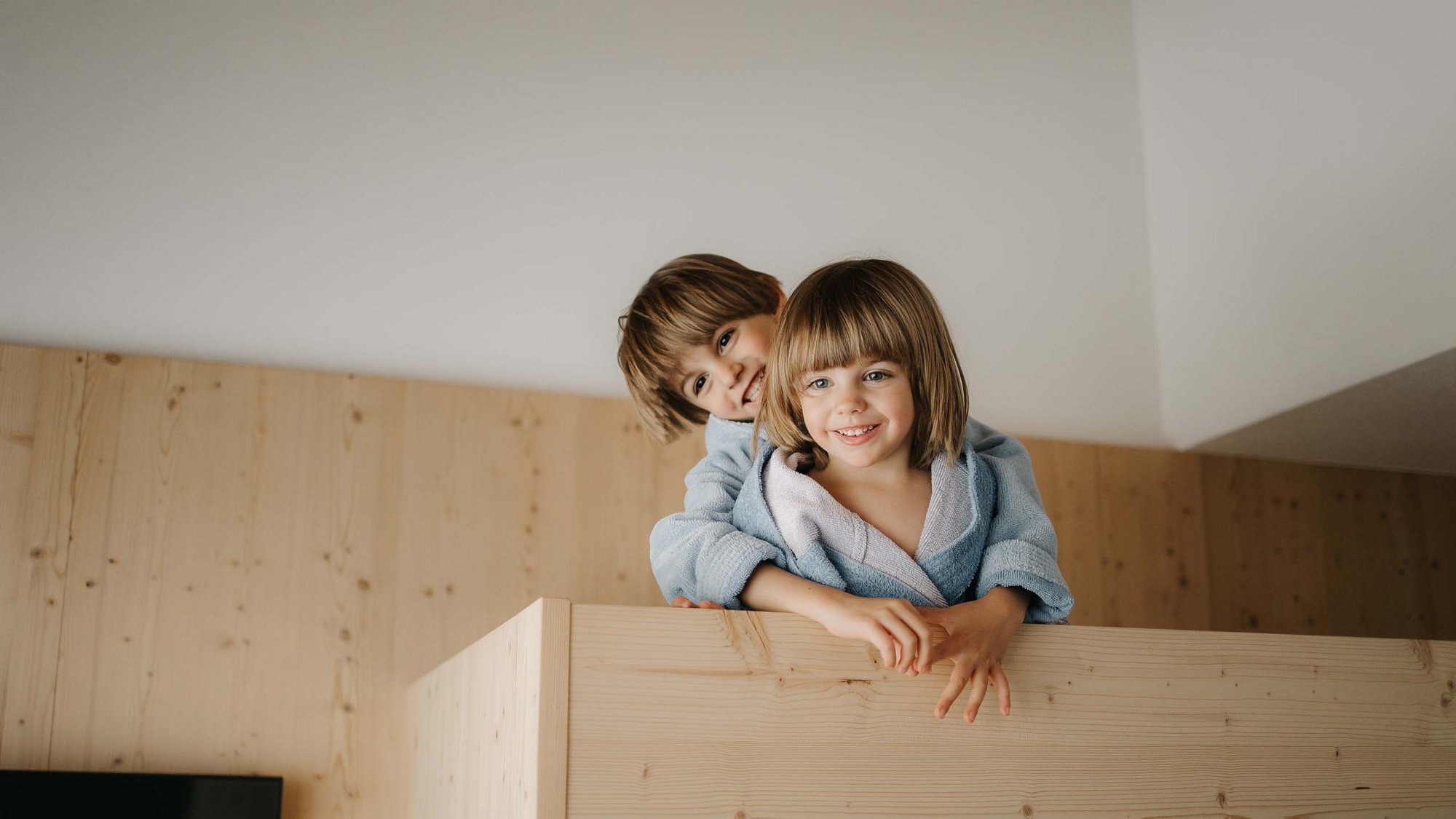 Family Resort Rainer: your accommodation in the Dolomites Two smiling children in bathrobes behind a wooden bed frame