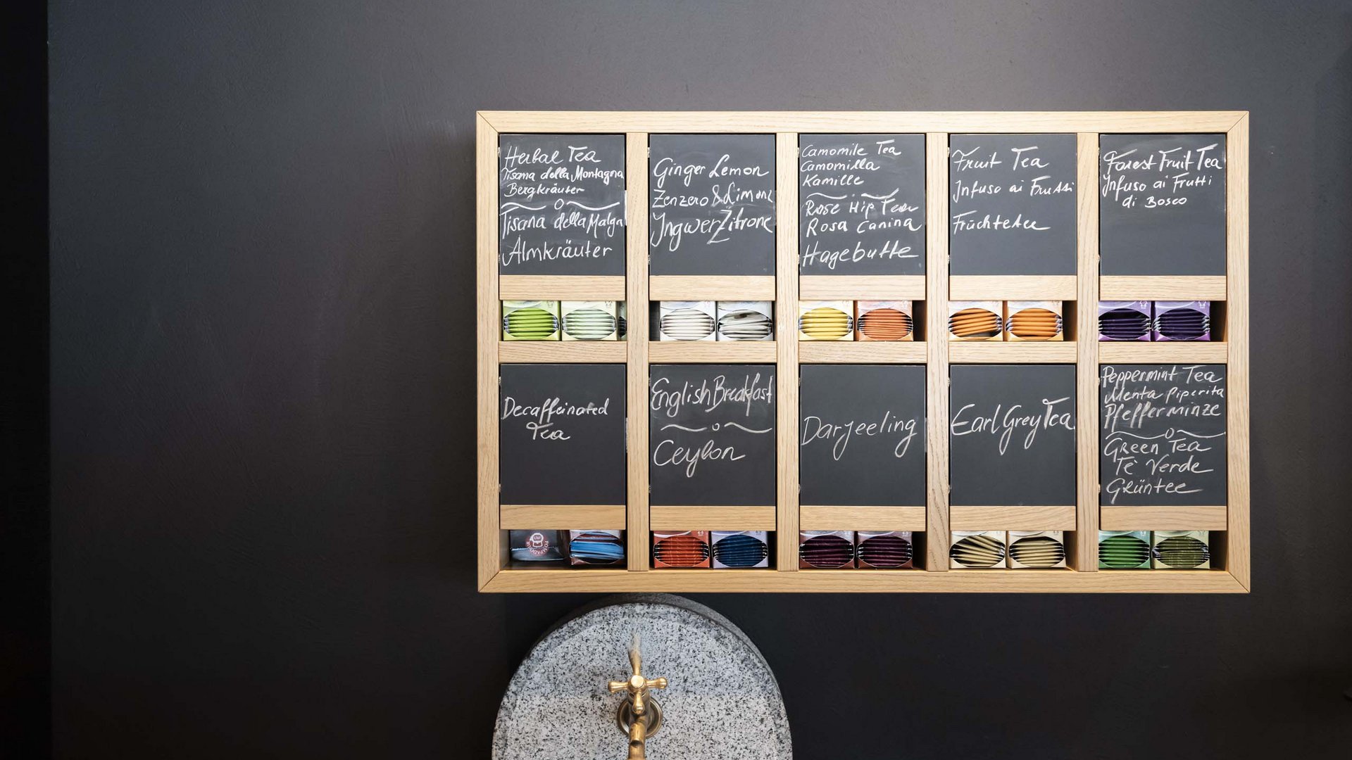 Family Resort Rainer Lavagna con tipi di tè e bustine di tè in scomparti su muro nero