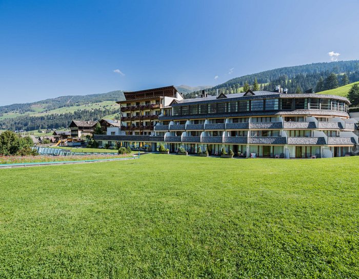 Ihr Familienresort in Südtirol Hotelgebäude mit Balkon und großem Rasen vor Berglandschaft unter blauem Himmel