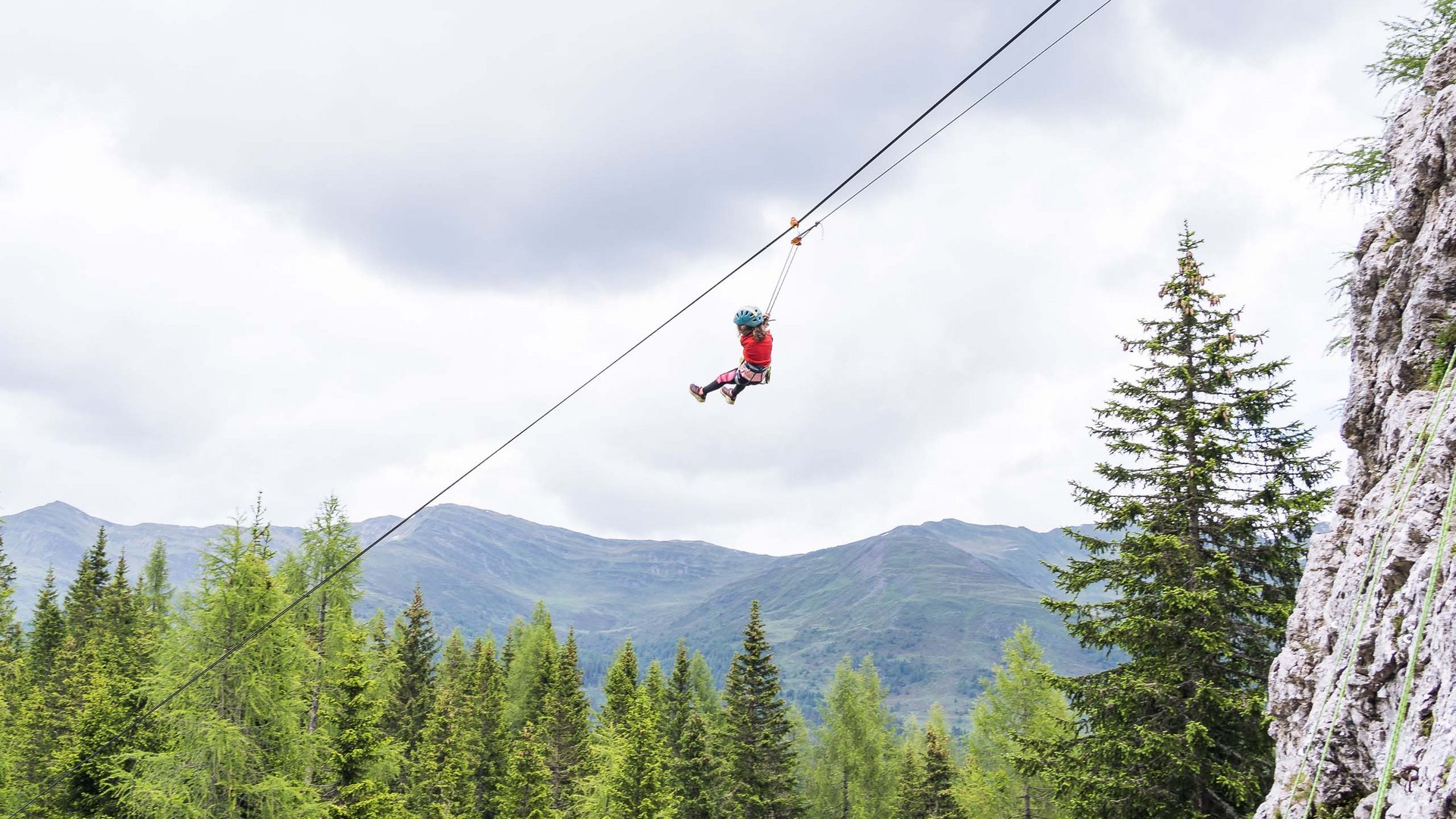 Qualche immagine del Rainer Bambino con casco su zipline sopra una foresta con montagne sullo sfondo