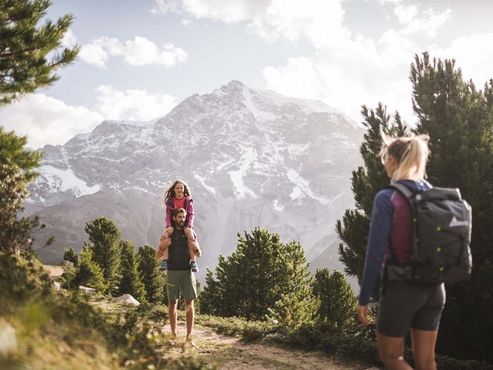 Man carrying child on shoulders hiking with woman in front of snowy mountains