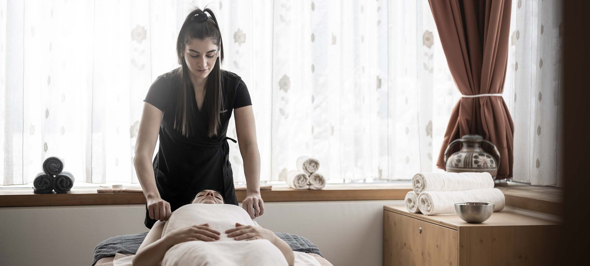 Relaxing wellness in the Dolomites: spa hotel in Sexten Woman receiving massage in bright room with white curtains and brown accents