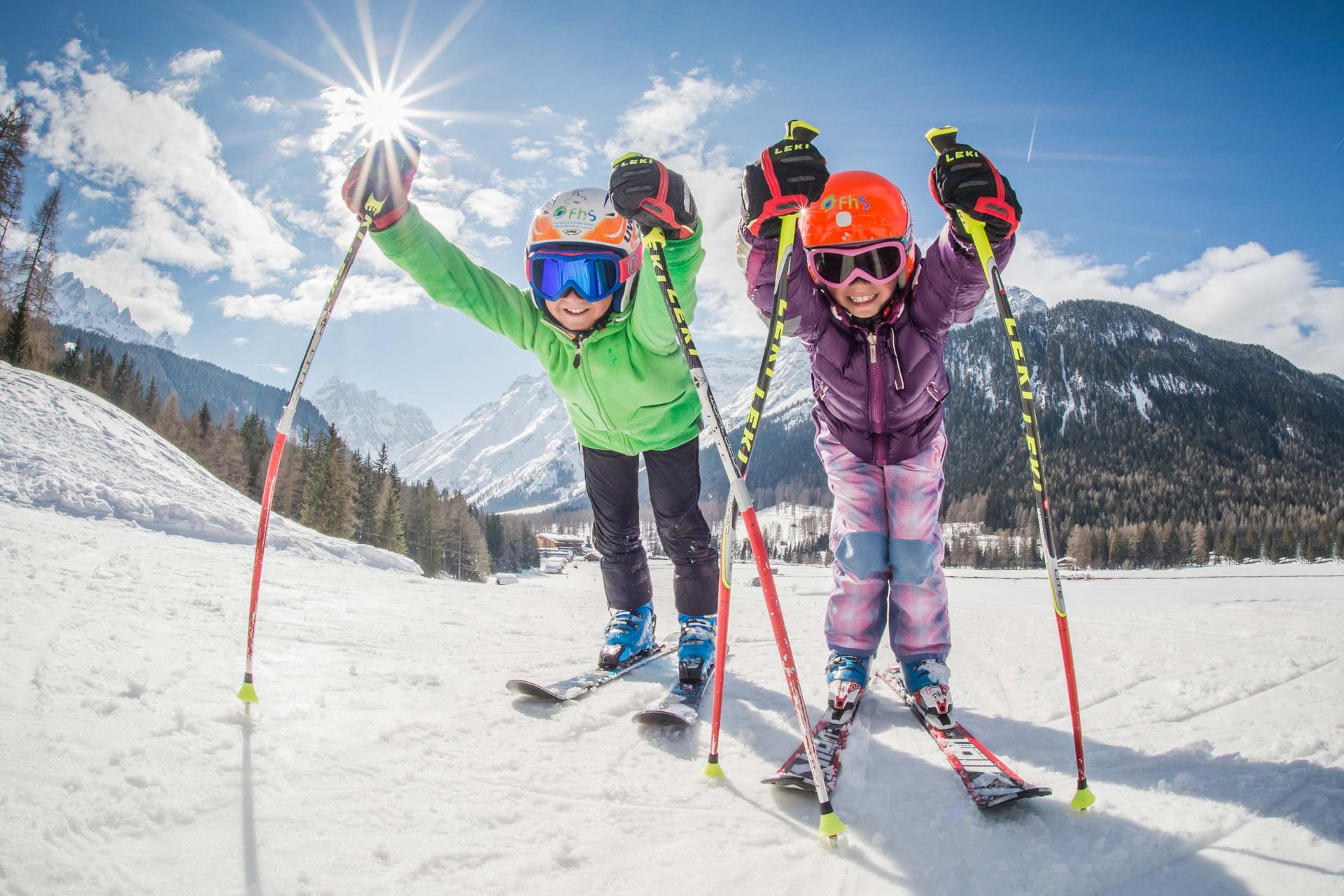 The in-house Rainer Ski School Two children in ski gear smiling on snow with mountains in the background.