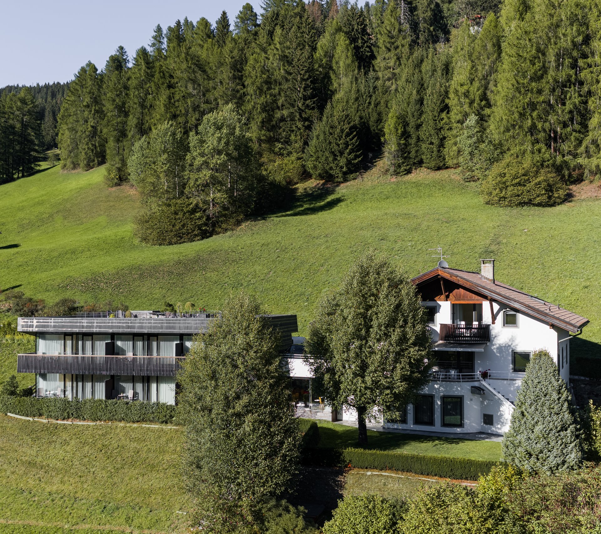 Haus und moderne Gebäude vor grünen Wiesen und gezackten Bergen bei klarem Himmel