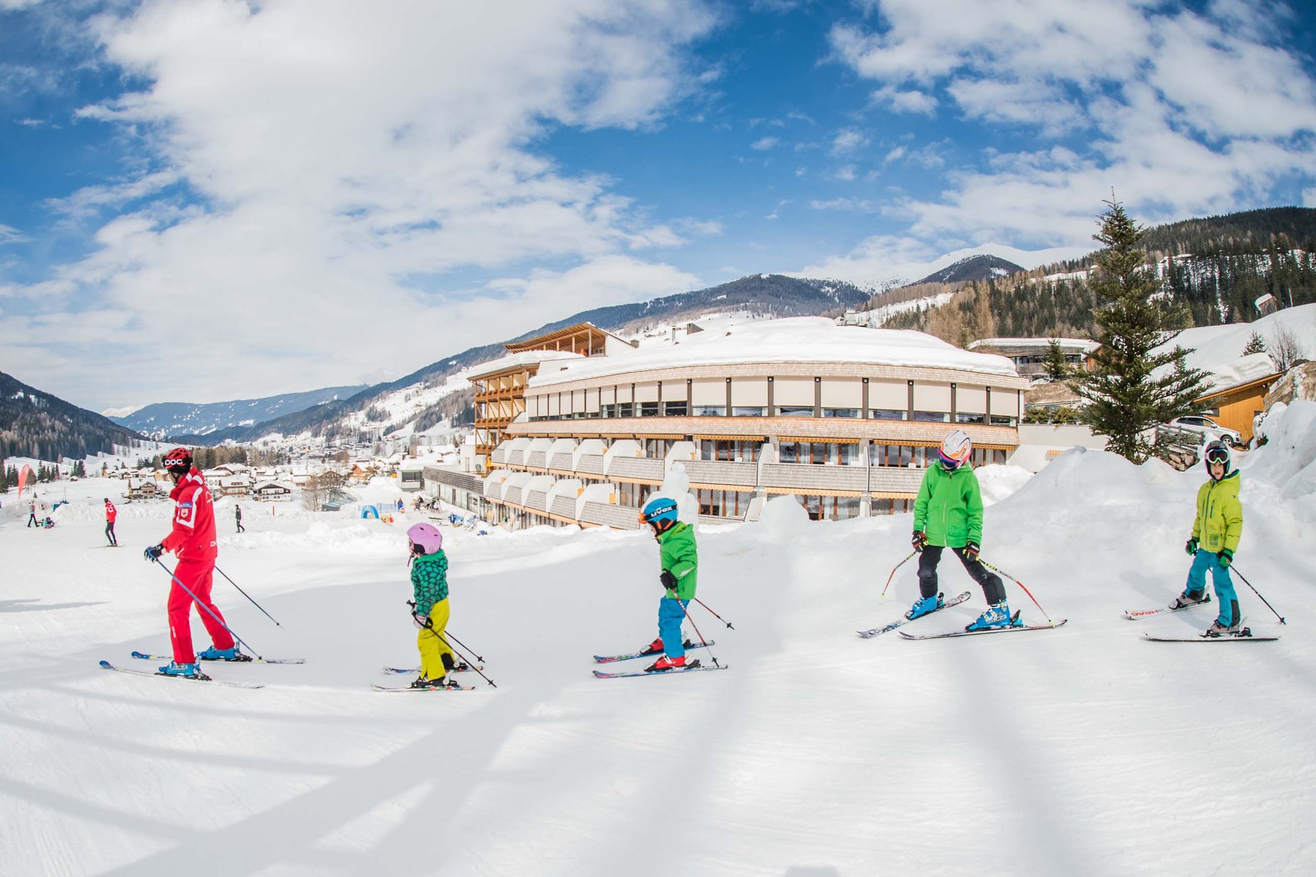 The in-house Rainer Ski School Ski instructor with children in ski lesson in front of hotel in snowy mountain landscape
