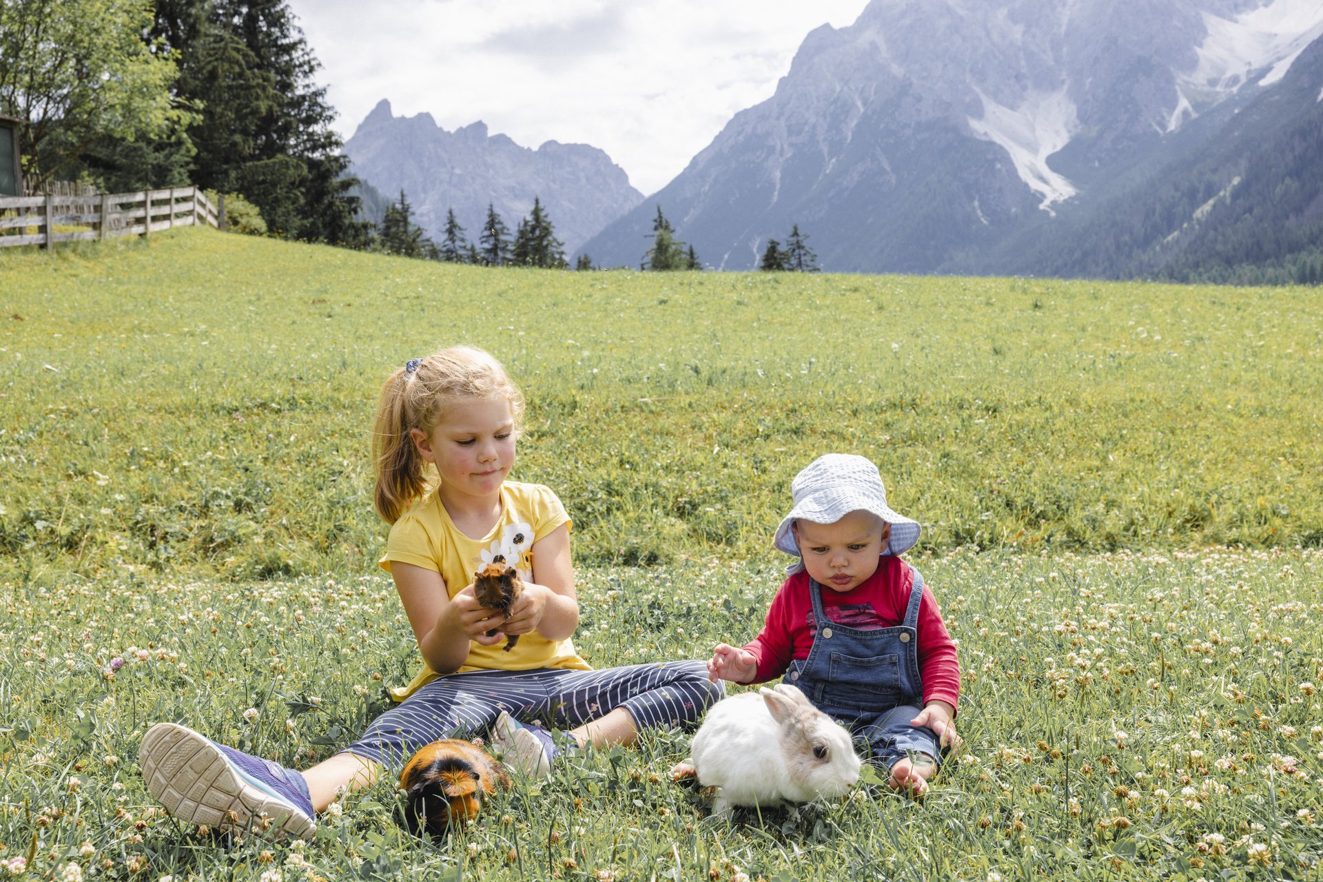 Il vostro family resort in Alto Adige Due bambini con un porcellino d’India e un coniglio seduti in un prato di montagna