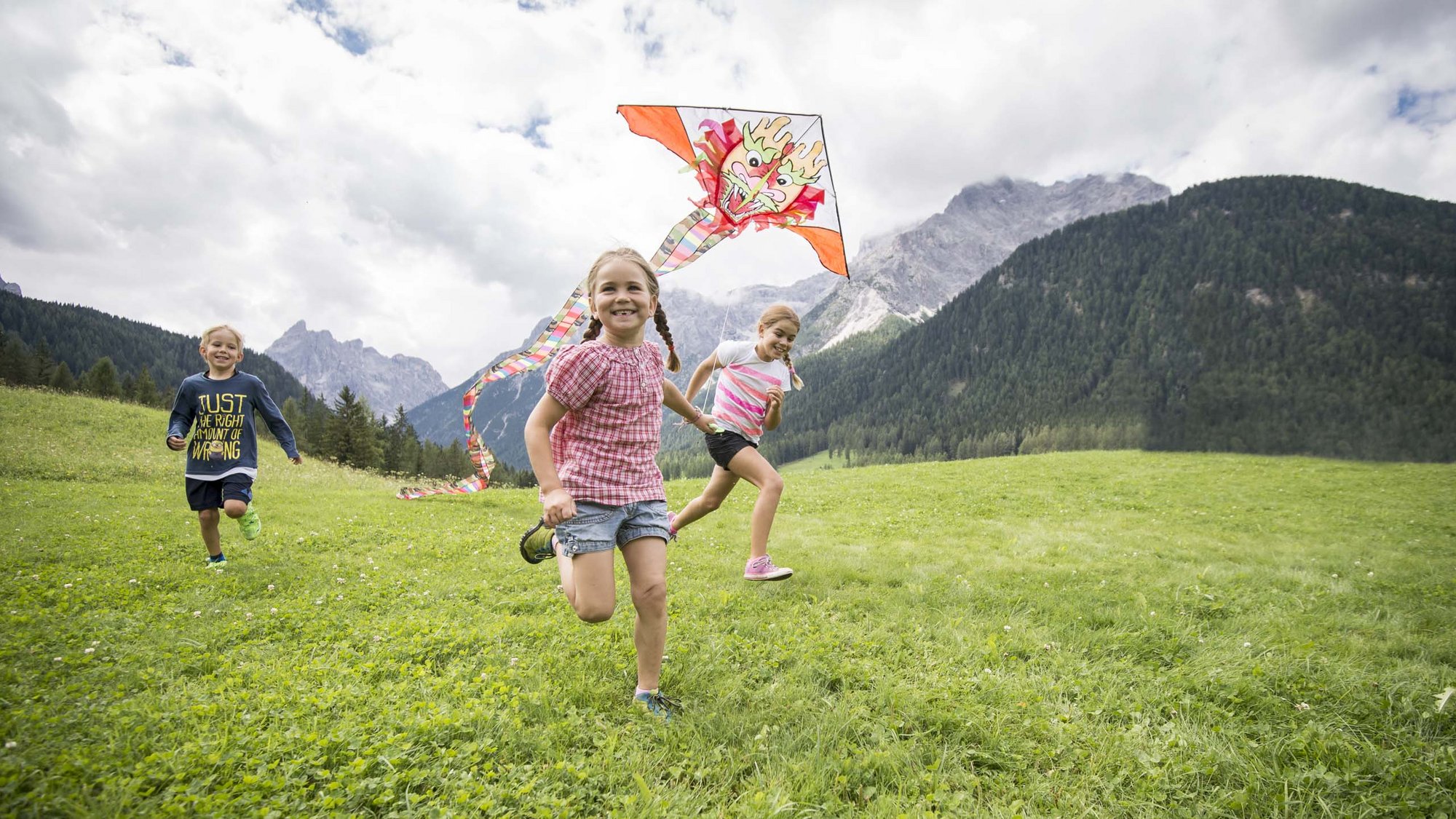 Stay at the Rainer: Family holidays in the Dolomites! Children running on a meadow flying a dragon-headed kite