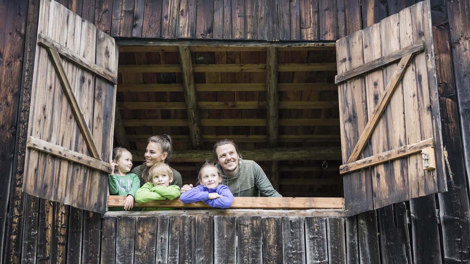 Stay at the Rainer: Family holidays in the Dolomites! Family looking out of open wooden window in rustic barn
