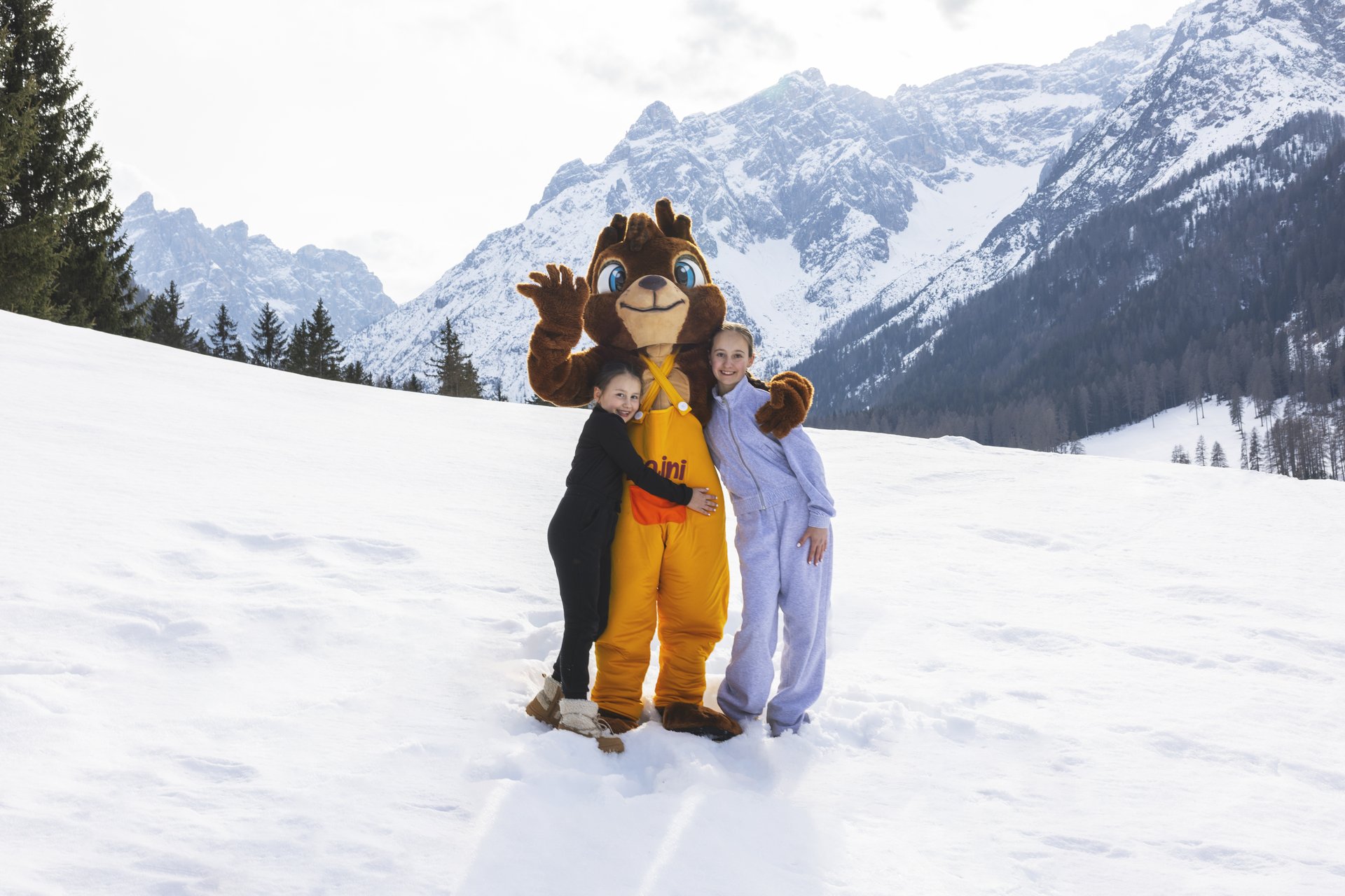 Il vostro family resort in Alto Adige Due bambine posano con una mascotte a forma di orso sulla neve, davanti a montagne innevate