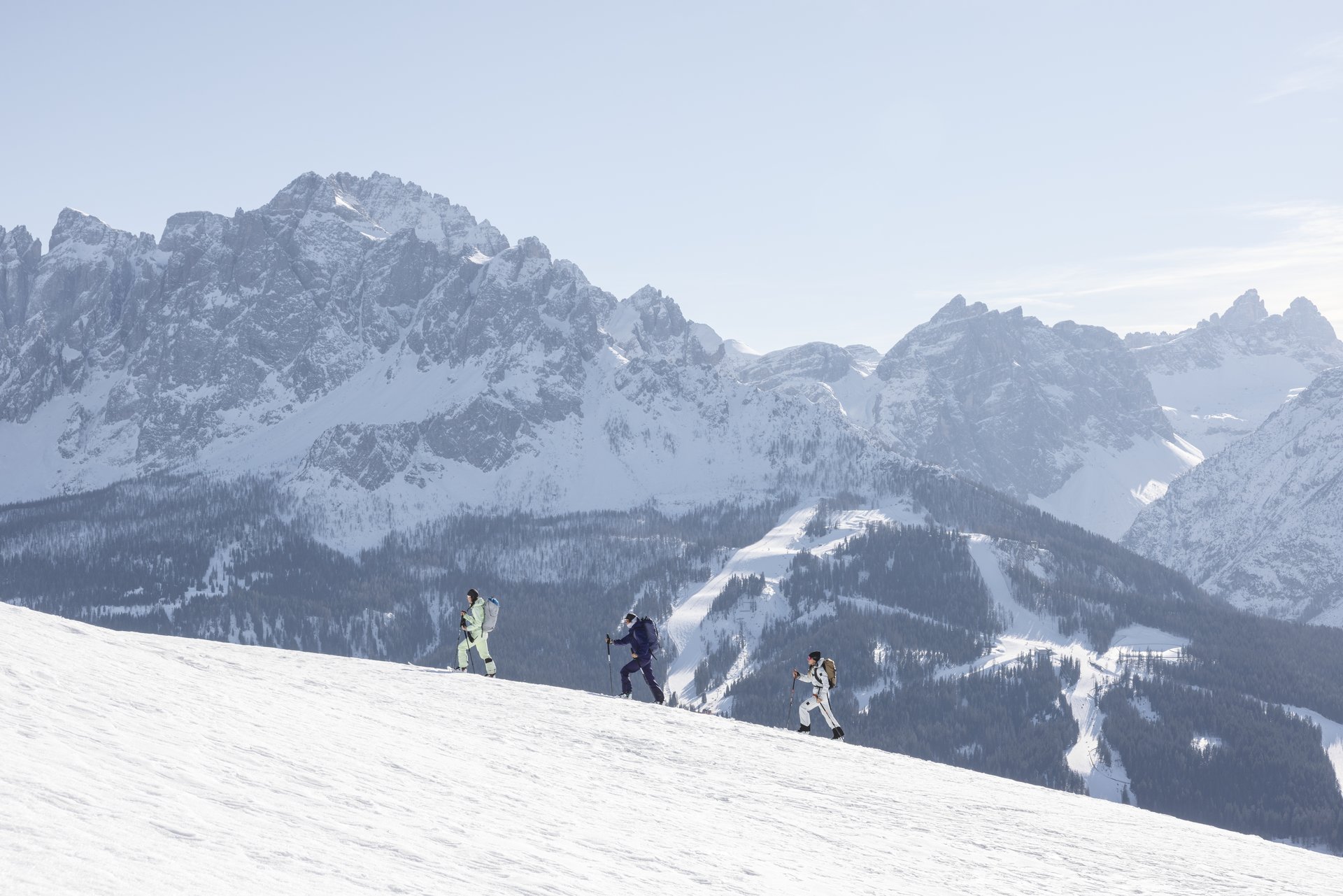 Drei Personen steigen mit Stöcken einen verschneiten Hang vor einer Bergkulisse hinauf