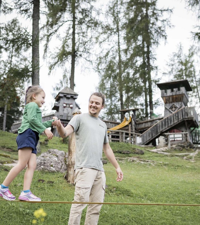 Your fulfilling family holiday Man helping girl on slackline in wooded park with playground in background