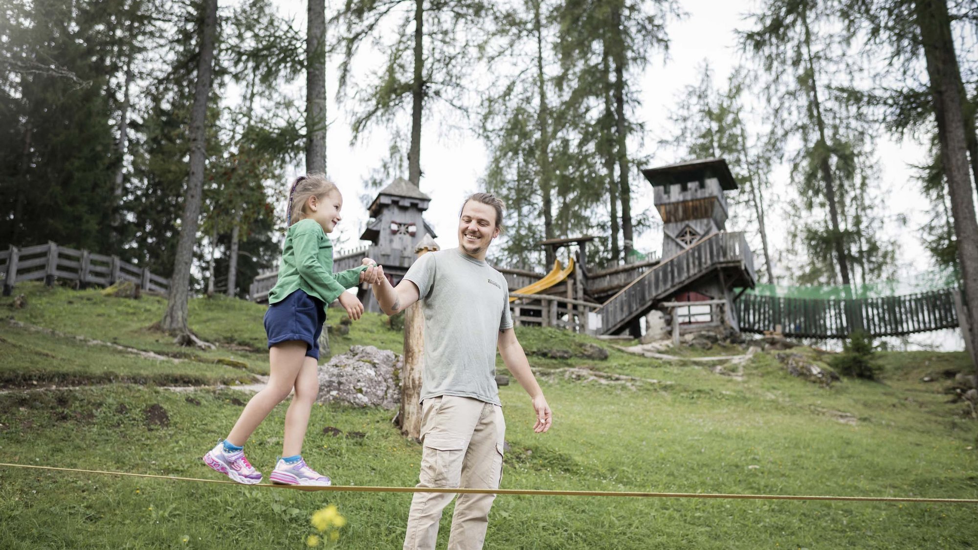Our family experiences Man helping girl on slackline in wooded park with playground in background