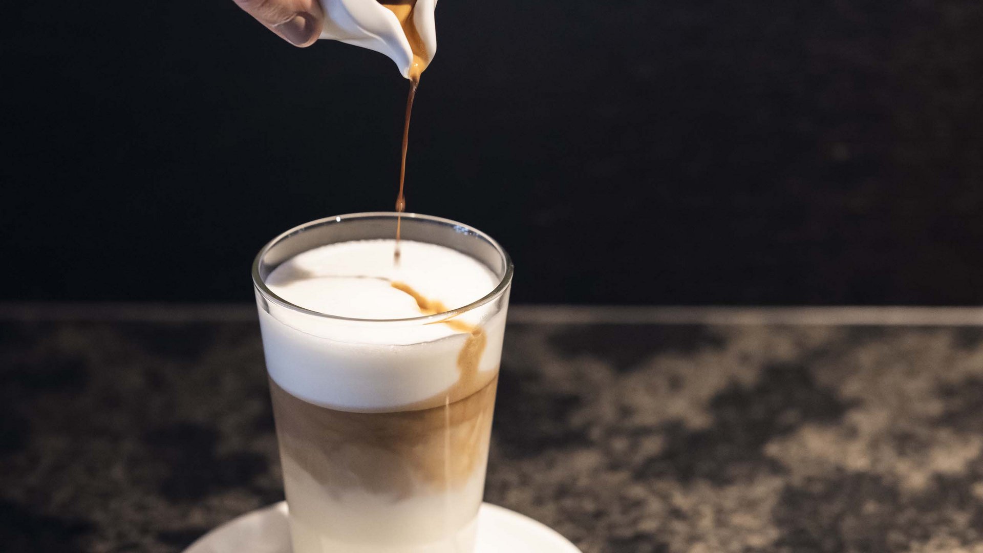 Family Resort Rainer Mano versa espresso in un bicchiere con schiuma di latte su piattino bianco