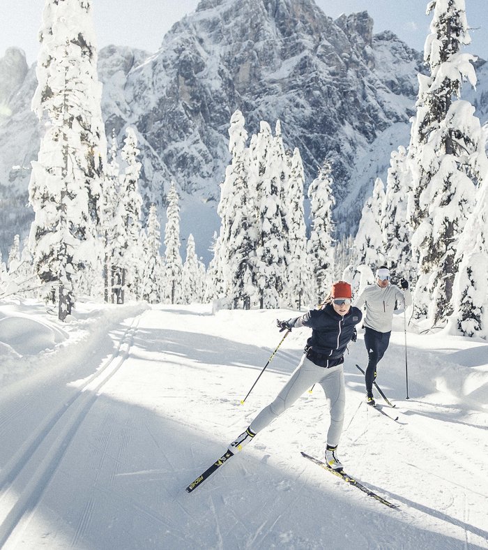 Our family experiences Two people cross-country skiing with snowy trees and mountains behind