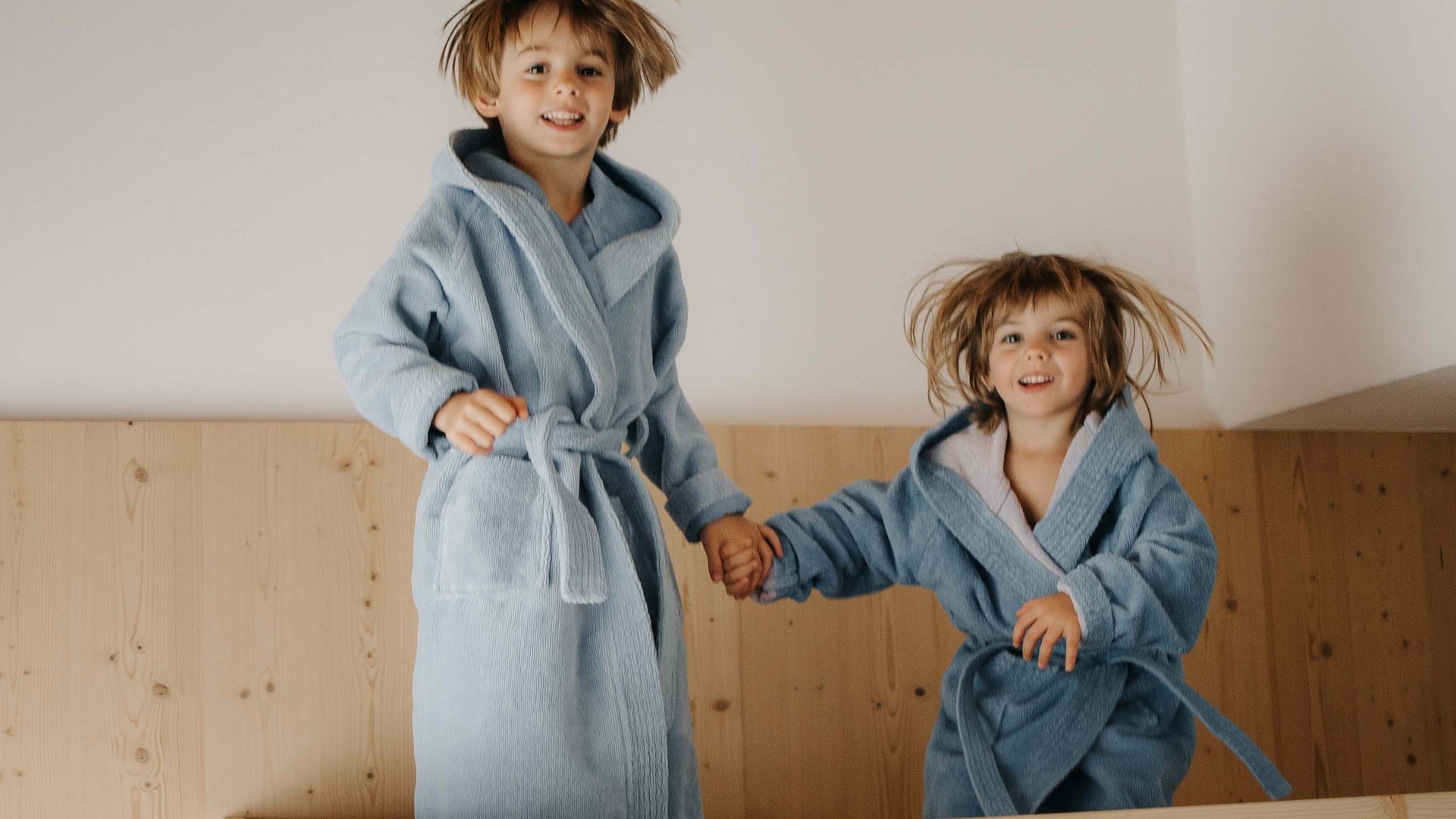 Family Resort Rainer Due bambini con accappatoi blu saltano felici su un letto di legno