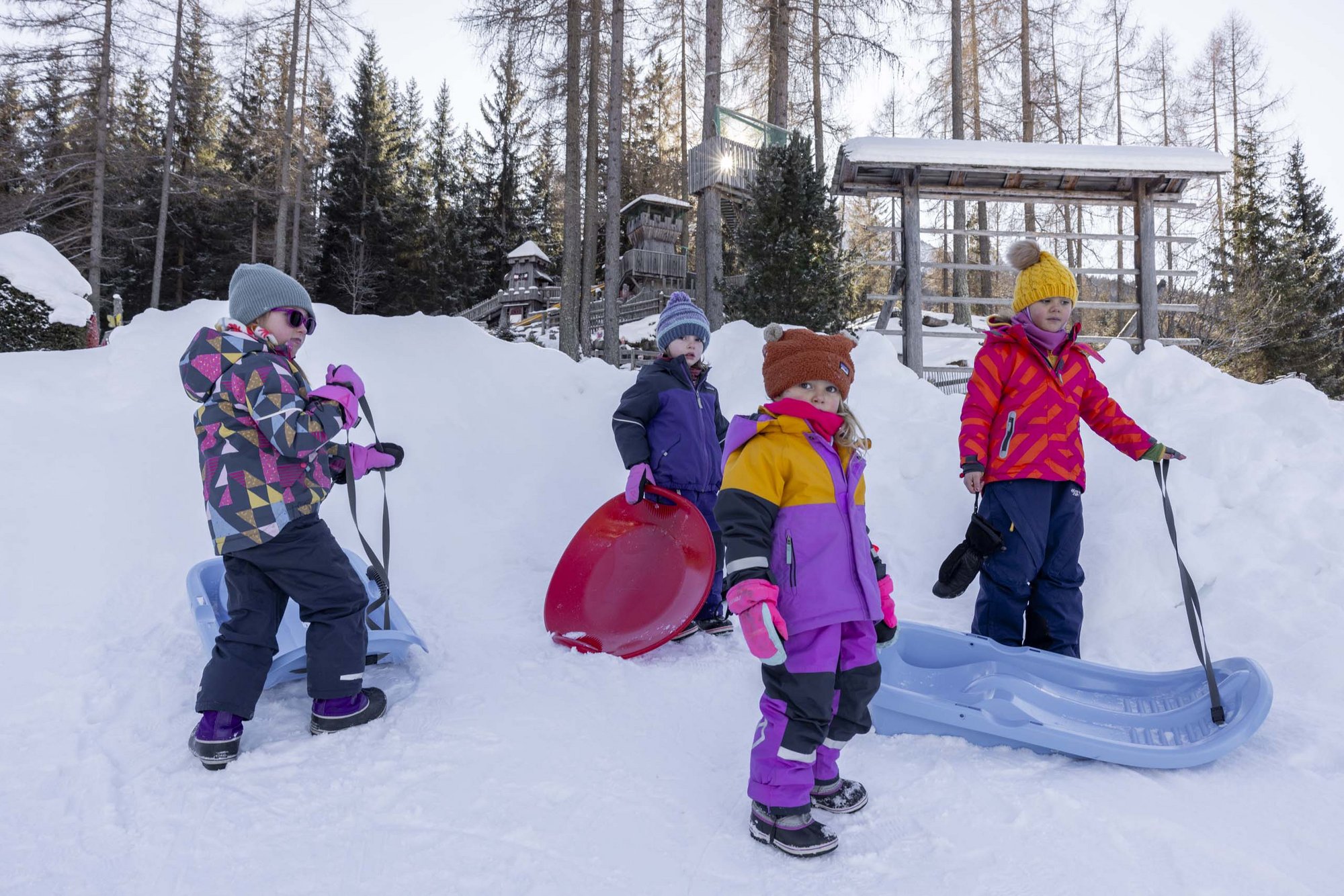 Stay at the Rainer: Family holidays in the Dolomites! Four children in colorful winter clothes with sleds in snowy forest