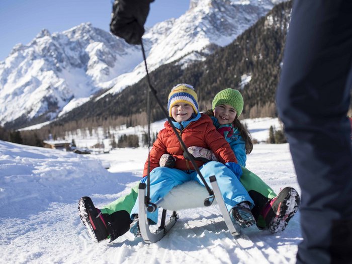 Your cross-country skiing hotel in the Dolomites Two happy children riding a sled in the snow with snowy mountains in the background