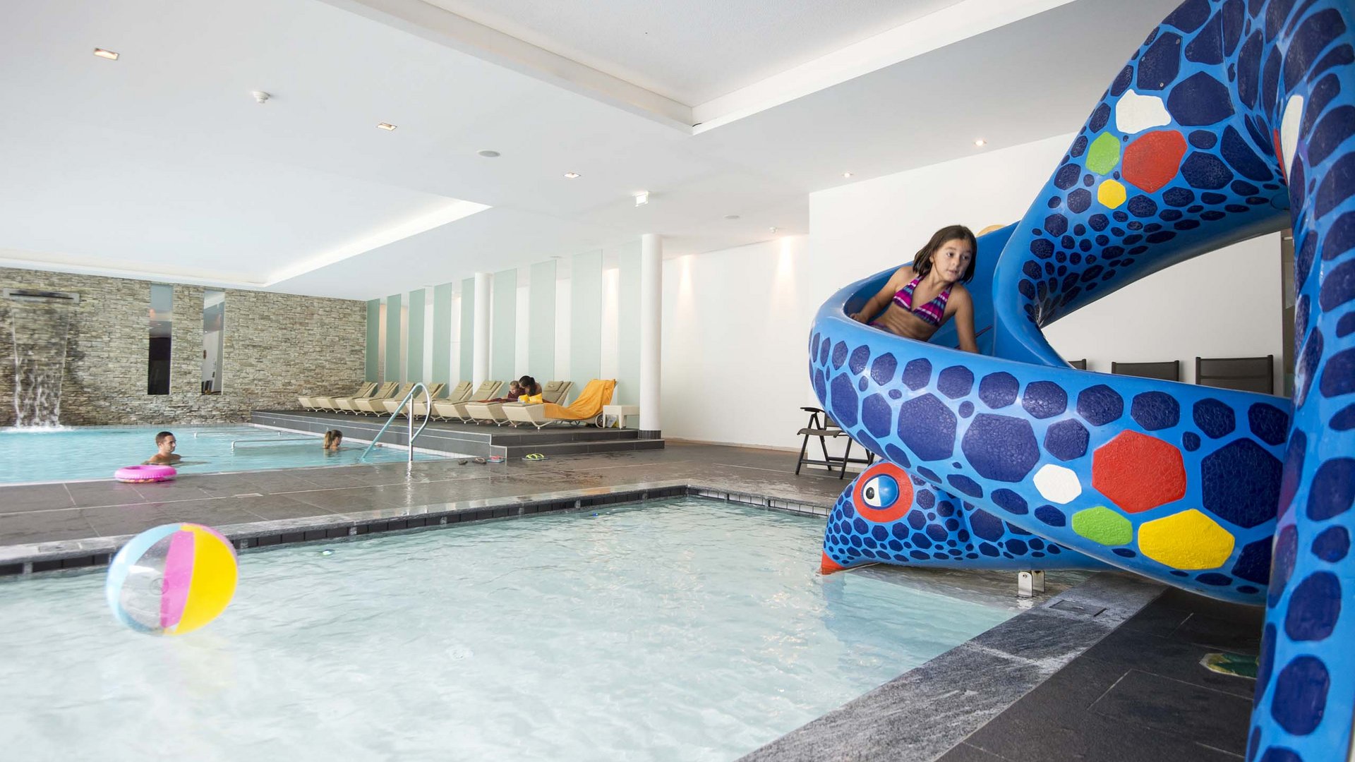 Family Resort Rainer Ragazza scivola su scivolo d'acqua blu in piscina coperta con bambini e lettini