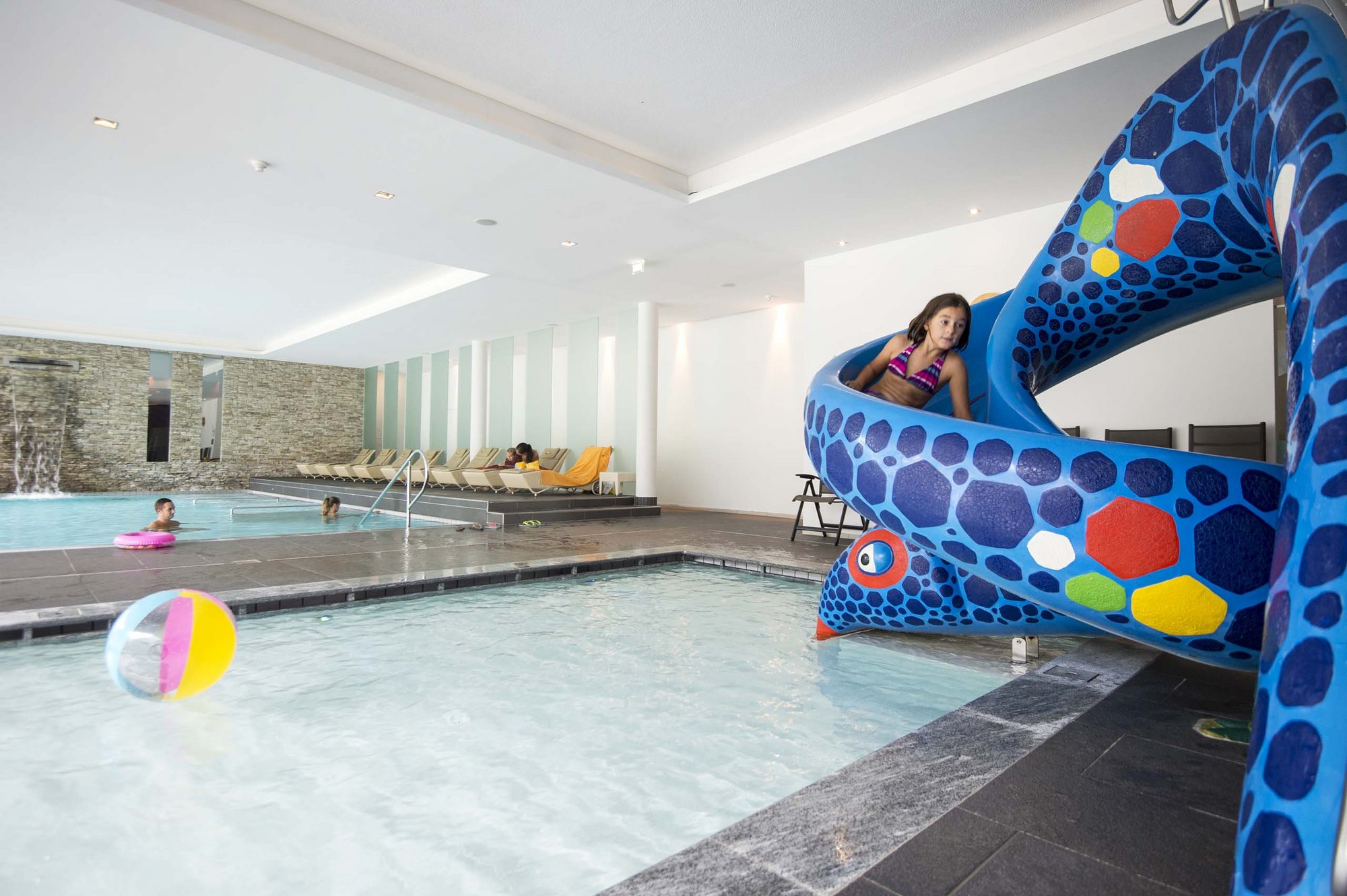 Ihr Familienresort in Südtirol Mädchen rutscht auf blauer Wasserrutsche ins Hallenbad mit Kindern und Liegestühlen