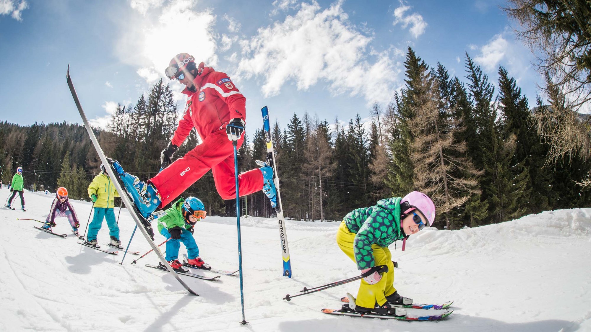 Stay at the Rainer: Family holidays in the Dolomites! Ski instructor jumps over children during skiing lesson on snowy sunny day