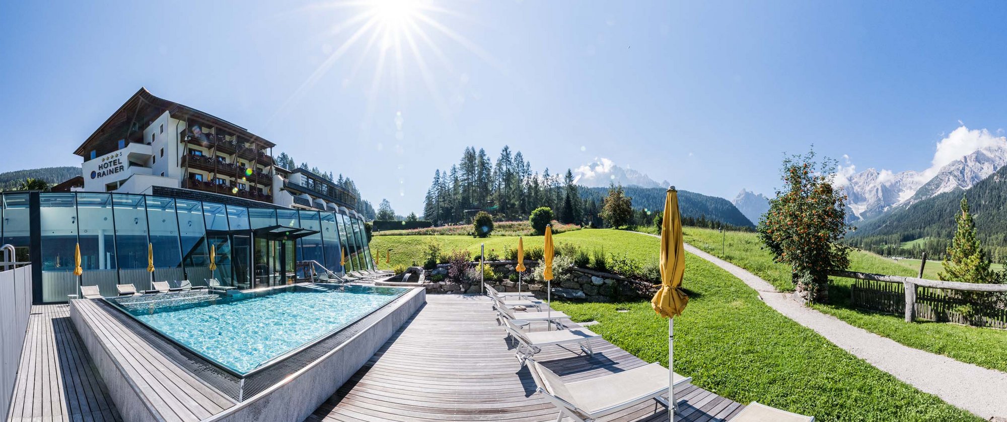 Your fulfilling family holiday Hotel Rainer with pool, lounge chairs, and mountain view on a sunny day