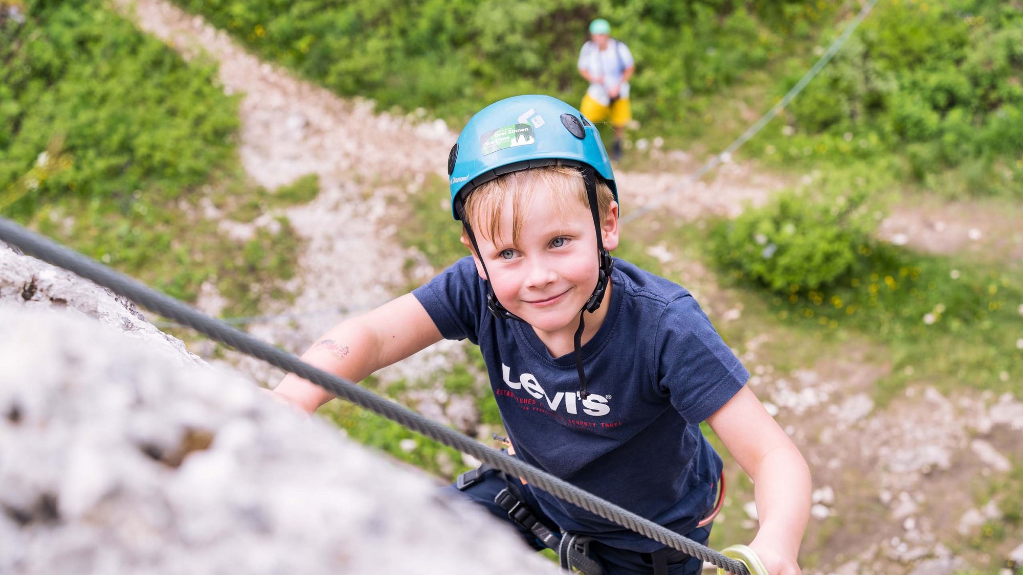 Stay at the Rainer: Family holidays in the Dolomites! Boy wearing helmet climbs rock face, person belays below on green terrain