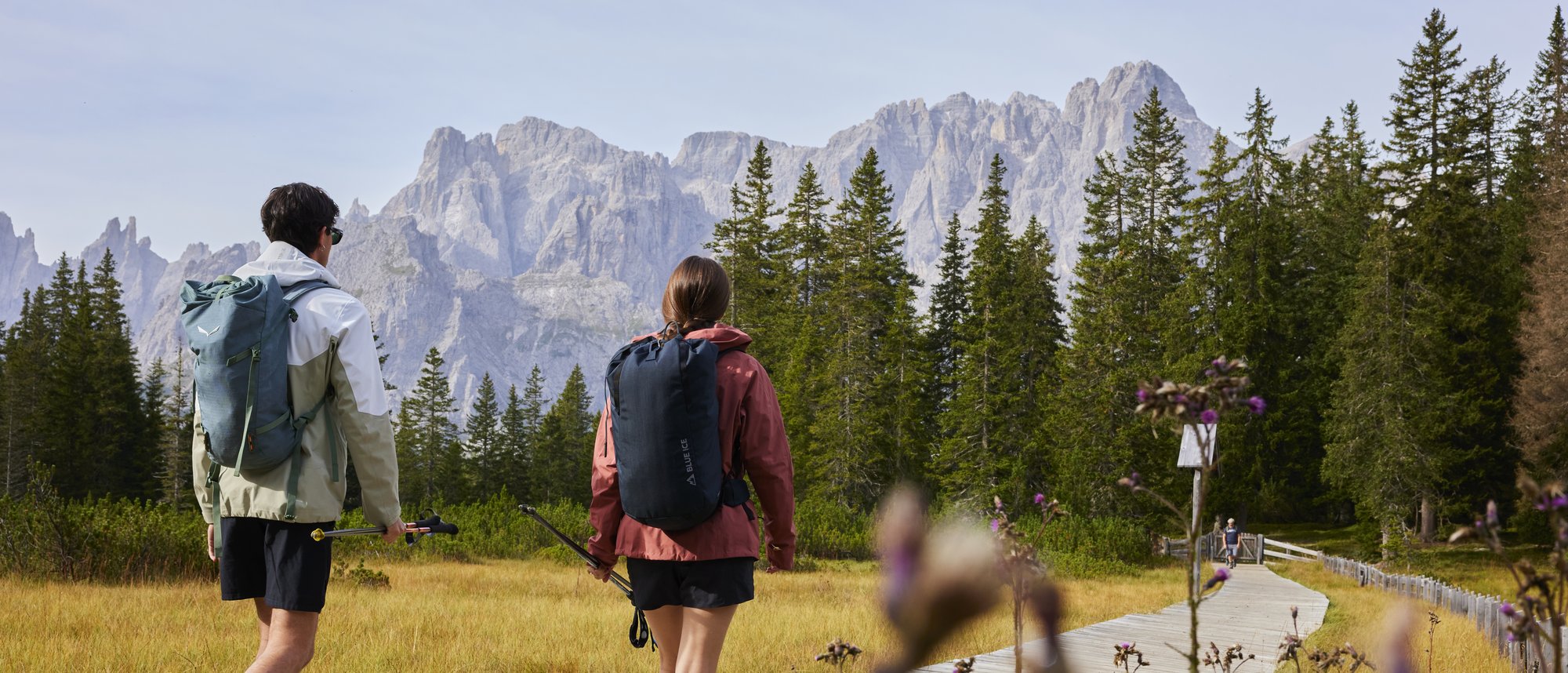 Kreuzbergpass - Alpe Nemes Zwei Wanderer mit Rucksäcken auf Holzsteg vor Bergkulisse und Nadelbäumen