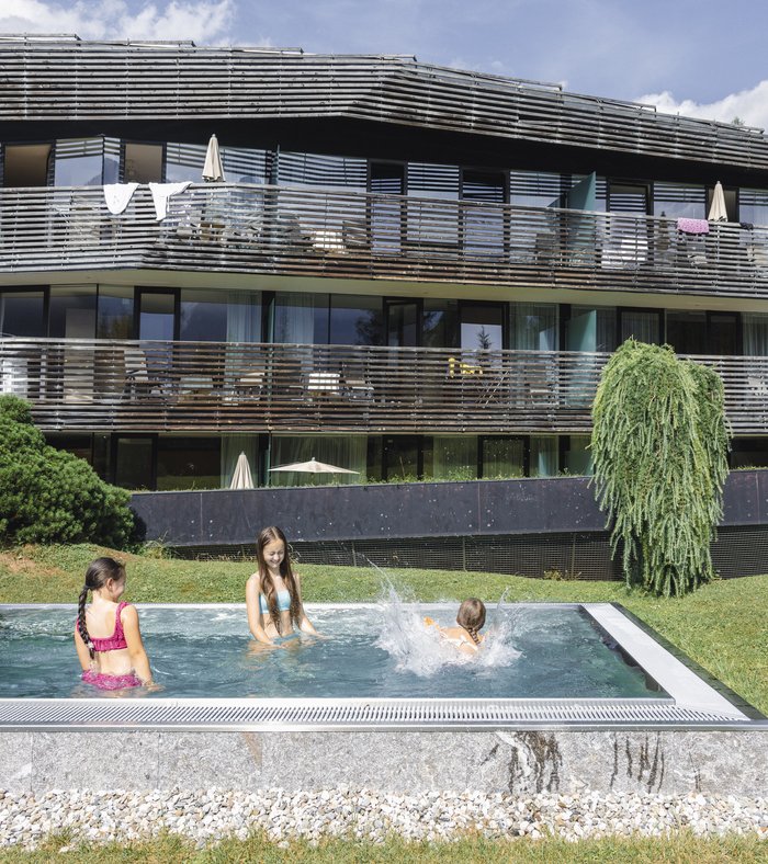 Il vostro family resort in Alto Adige Tre bambini fanno il bagno nella piscina all’aperto davanti a un edificio moderno su un pendio verde