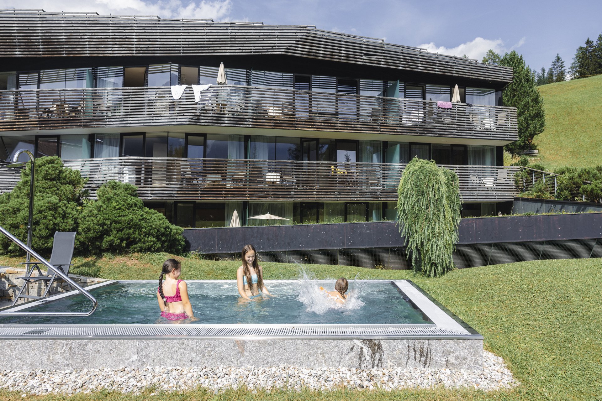 Il vostro family resort in Alto Adige Tre bambini fanno il bagno nella piscina all’aperto davanti a un edificio moderno su un pendio verde
