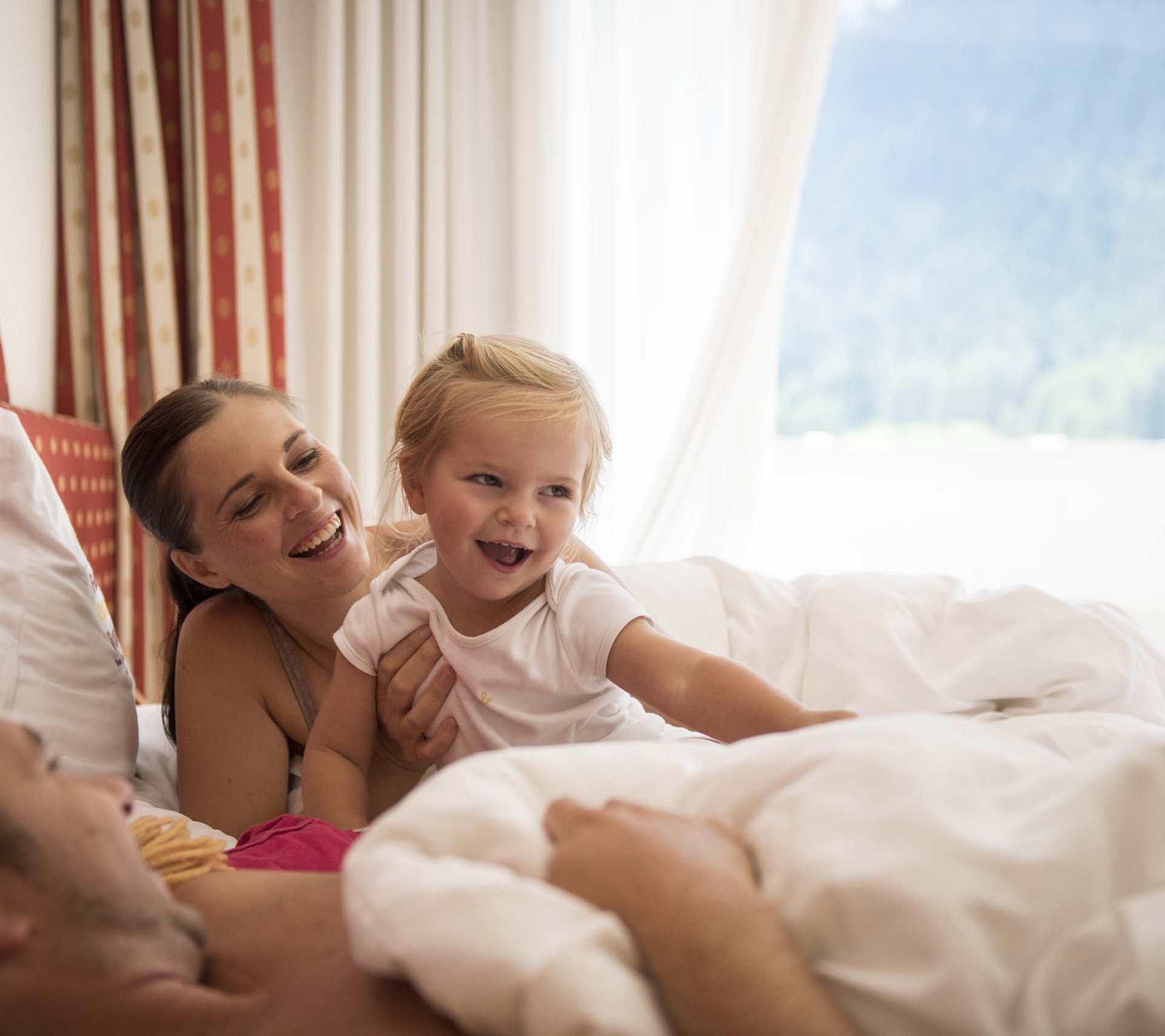 Property detail page - Hotel Rainer Happy family with child playing in bed in bright, sunny bedroom