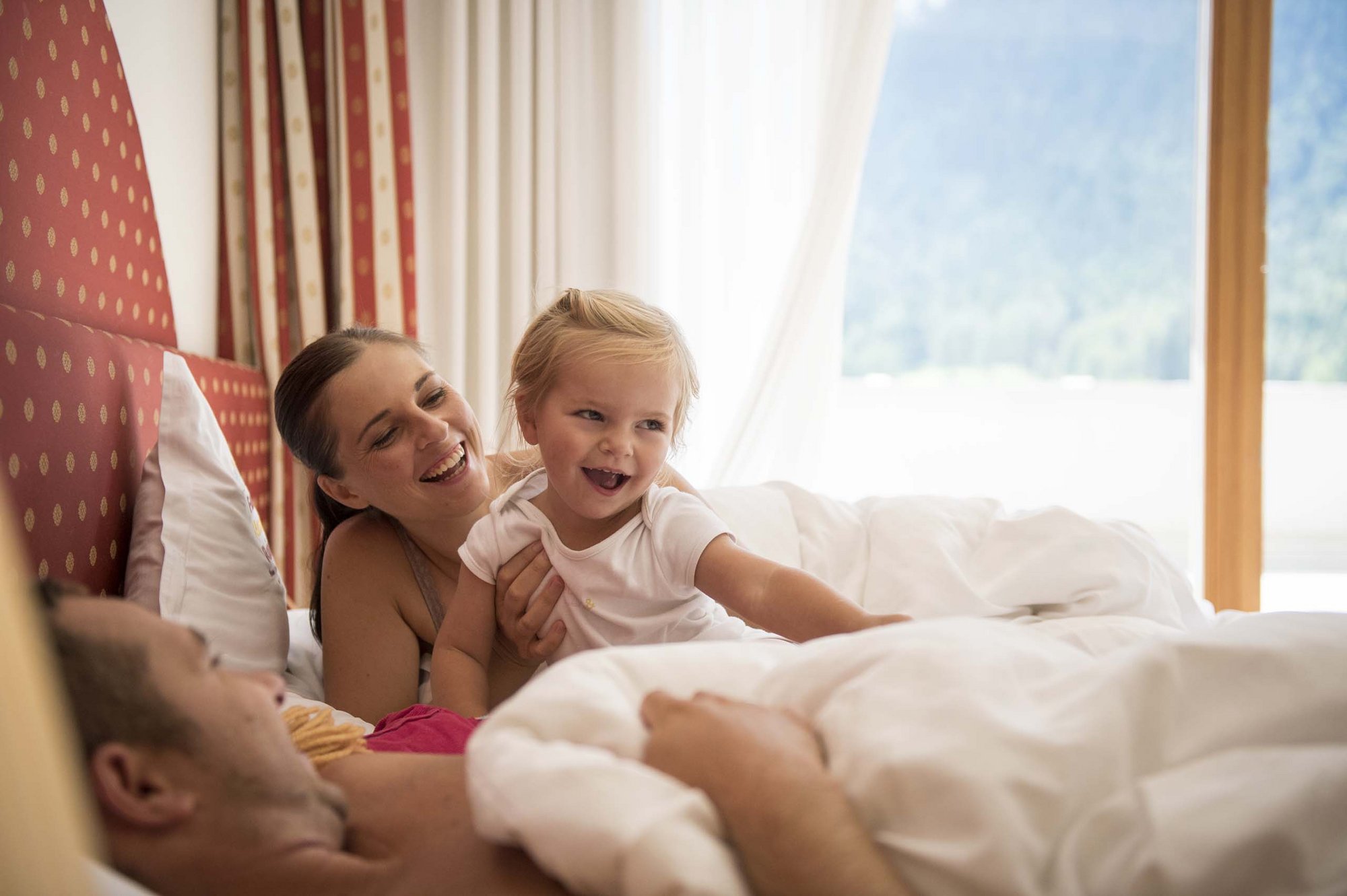 Family Resort Rainer: your accommodation in the Dolomites Happy family with child playing in bed in bright, sunny bedroom