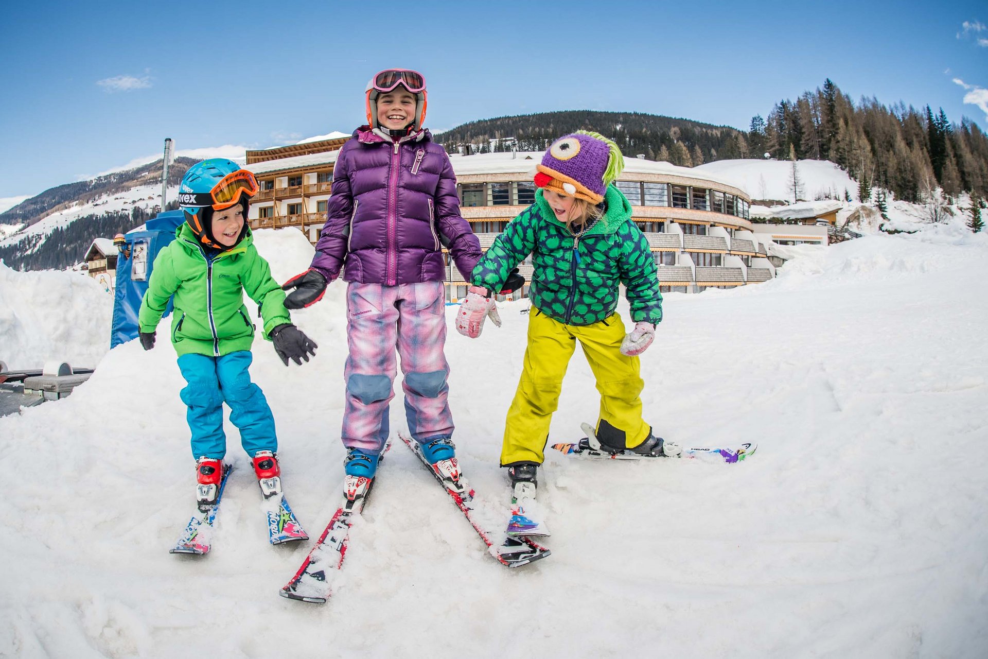 The in-house Rainer Ski School Three children in colorful ski gear playing in the snow in front of a hotel