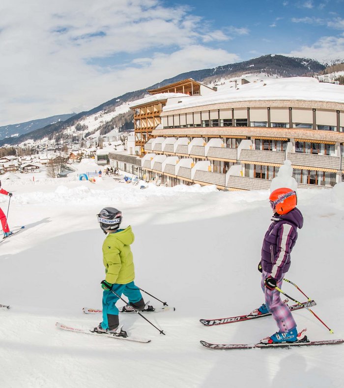 Ihr Familienresort in Südtirol Kinder lernen Skifahren vor einem Hotel im verschneiten Bergdorf