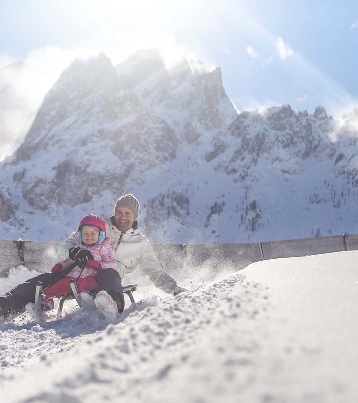 Stay at the Rainer: Family holidays in the Dolomites! Father and child sledding happily in snow with snowy mountains in background