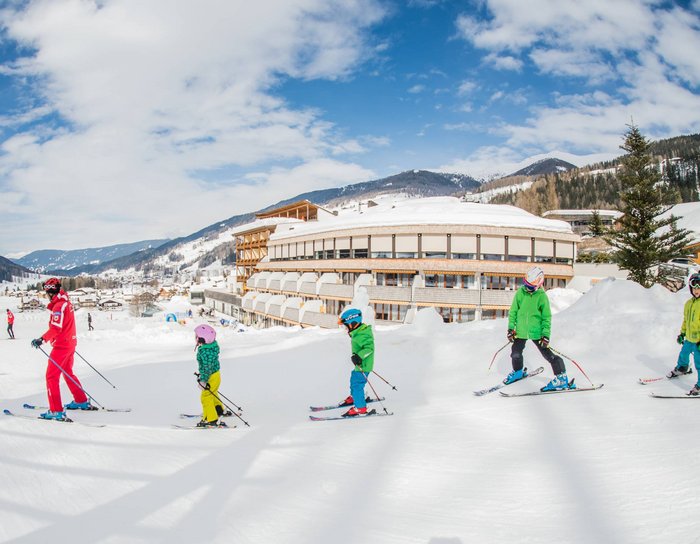 Ihr Familienresort in Südtirol Skikurs mit Kindern und Lehrer vor einem Hotel in verschneiter Berglandschaft