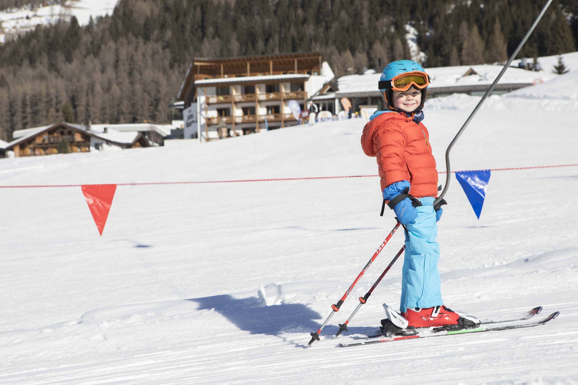 The in-house Rainer Ski School Child wearing helmet and skis standing on snow with mountain hotel in background