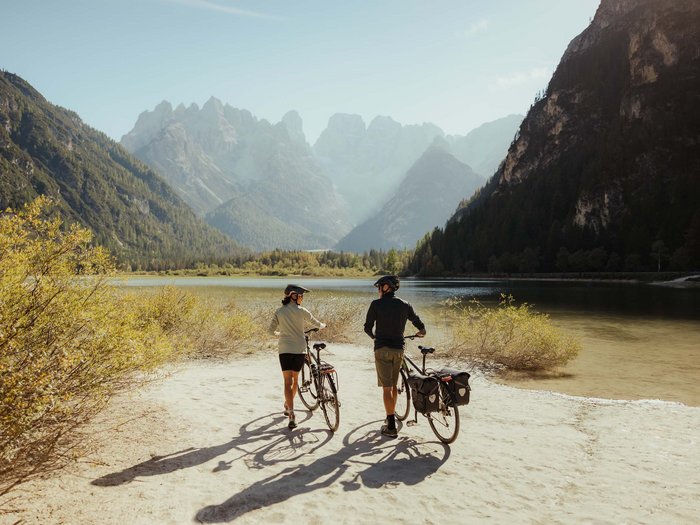 Sexten: a paradise for mountain lovers Two cyclists walking with bikes along a lake surrounded by mountains