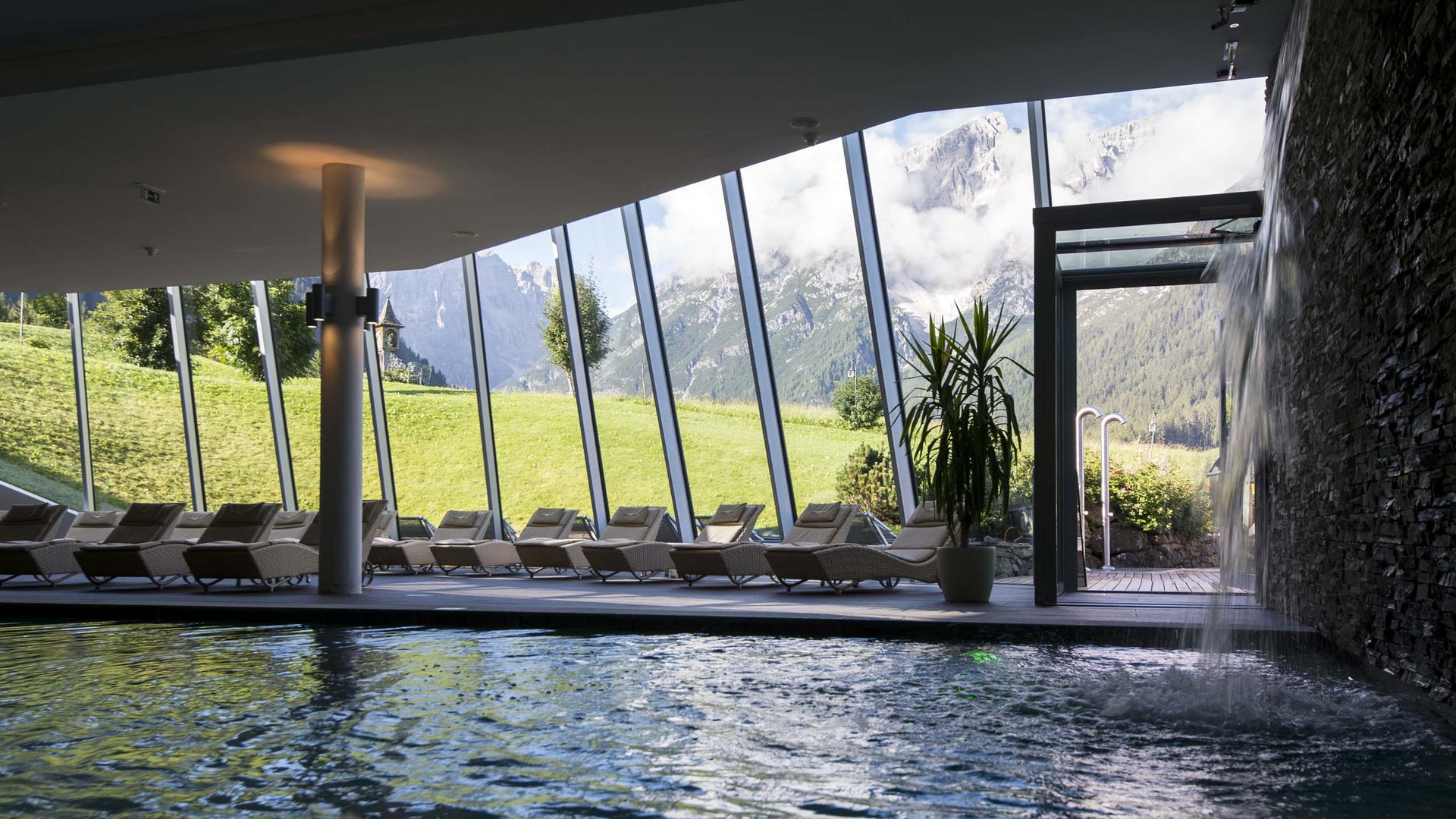 Family Resort Rainer Piscina interna con cascata e lettini davanti a grandi vetrate con vista sulle montagne