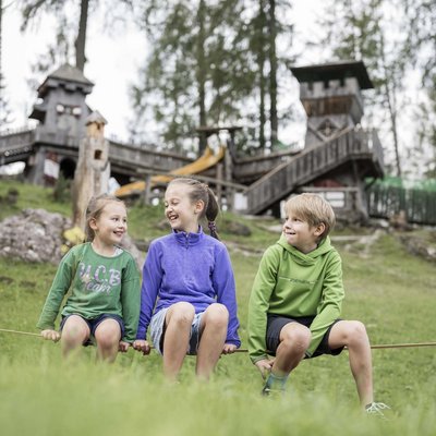 Ihr Familienresort in Südtirol Drei lachende Kinder sitzen auf einem Seil vor einem Holzspielplatz im Grünen