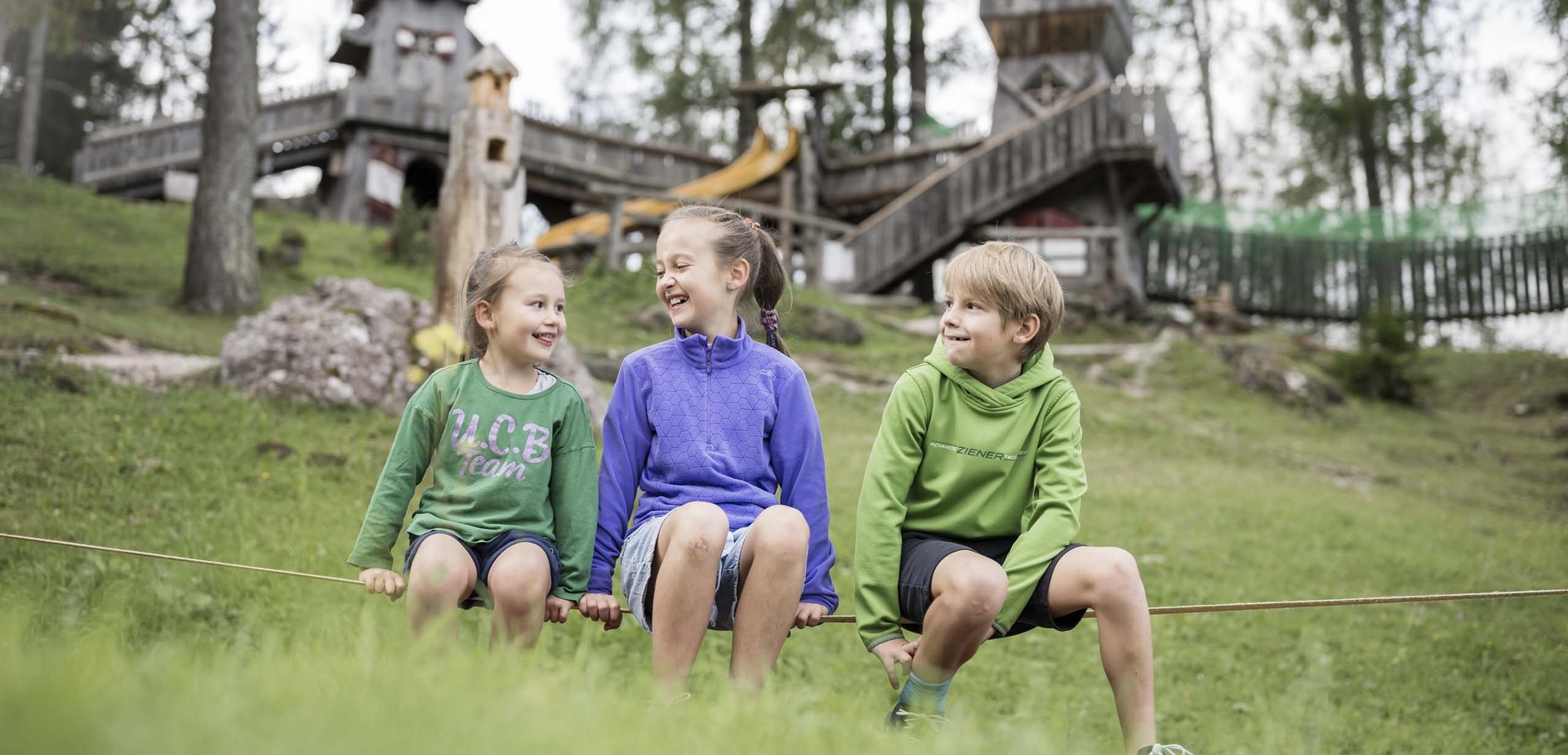 Useful information about your holiday Three smiling children sitting on a rope in front of a wooden playground outdoors