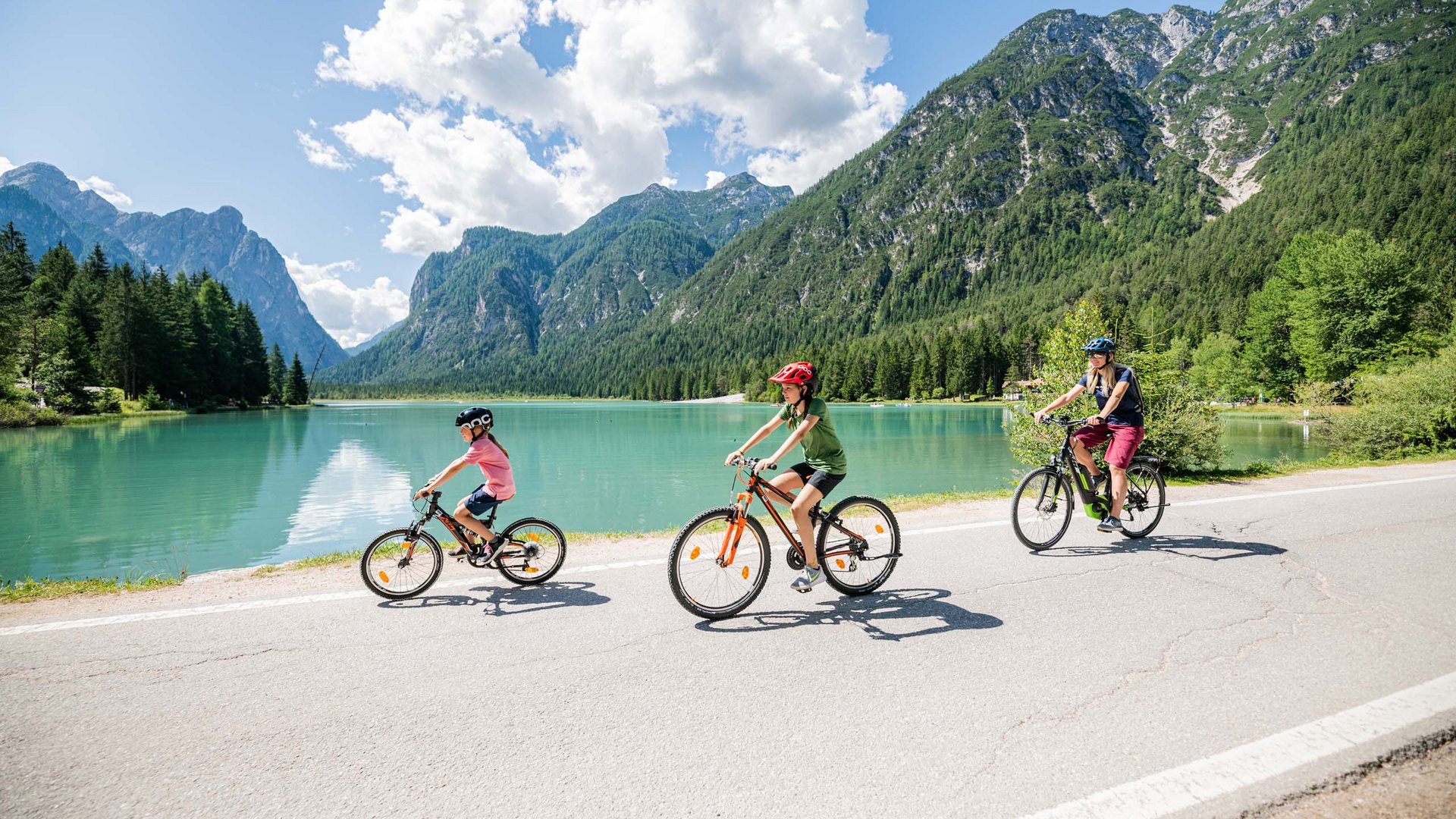 Qualche immagine del Rainer Tre persone in bicicletta vicino a un lago circondato da montagne in una giornata soleggiata